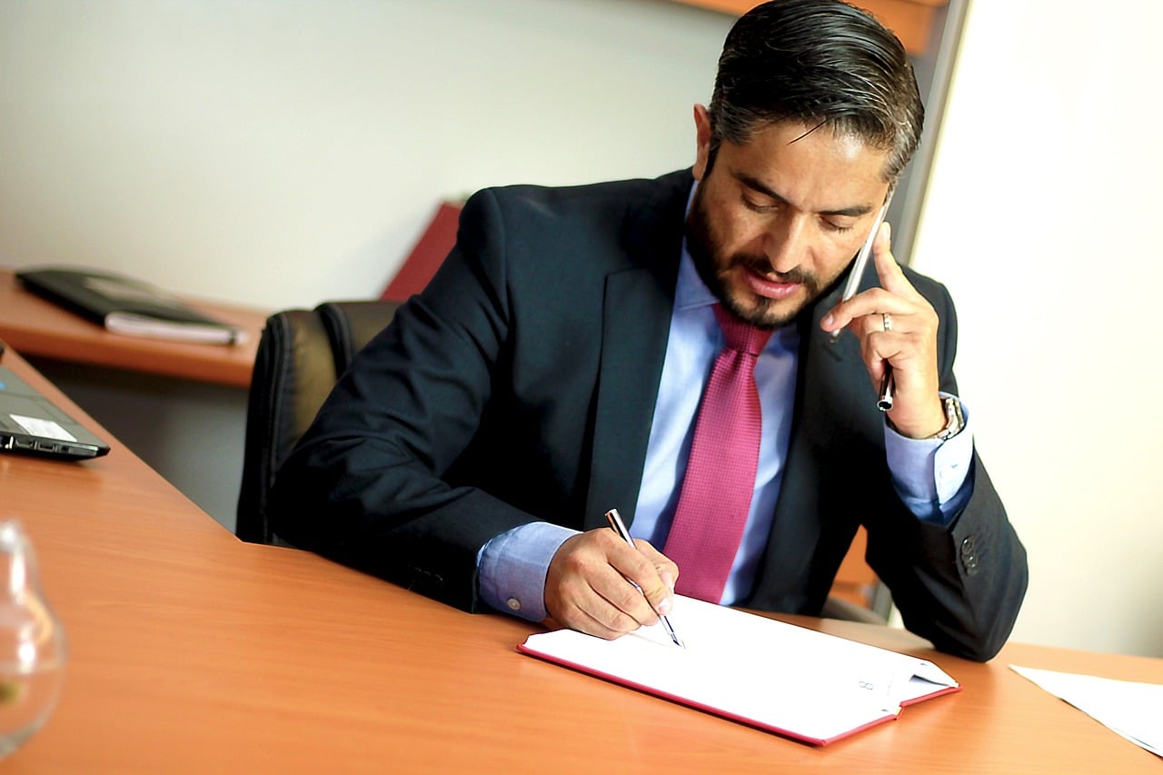 Un homme en costume-cravate est assis à un bureau, parlant au téléphone tout en écrivant dans un cahier.