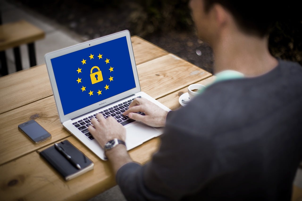 Une personne utilise un ordinateur portable affichant le drapeau de l'Union européenne avec un symbole de cadenas, indiquant la sécurité des données, alors qu'elle est assise à une table extérieure en bois avec un cahier, un stylo et une tasse de café.